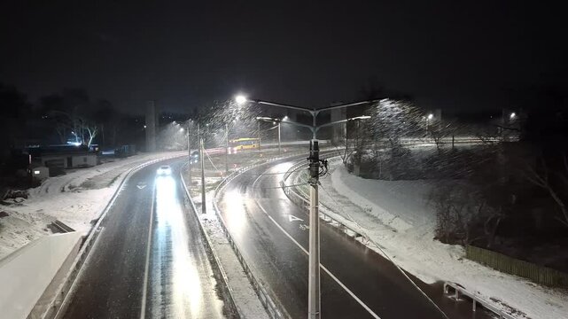 Winter, snow is falling. Lanterns on poles shine on the winter road. Cars and yellow bus drive on the slippery road 