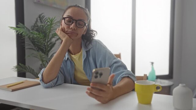 Woman with glasses resting chin on hand while scrolling smartphone at a table with yellow mug and notebook in a bright room in building; boredom distraction.