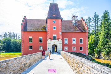 Fototapeta premium Visitors walk across a stone bridge to Cervena Lhota Castle. The bright red building stands out against green trees. It is a sunny day, and the water below reflects the castle.