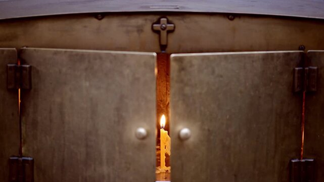 A candle burns softly, casting warm light through the gap of heavy metal doors