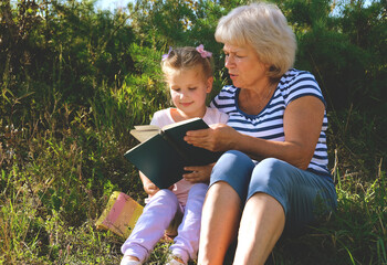 Fototapeta premium Happy grandmother and granddaughter reading book of stories or novel at leisure while sitting in the garden, backyard. Grandmother delighting in storytelling, engaged in reading a book