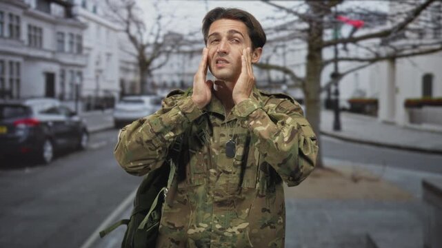 Soldier man in camouflage with dog tags presses temples with both hands on a street lined with parked cars and white buildings for headache; stress homecoming.