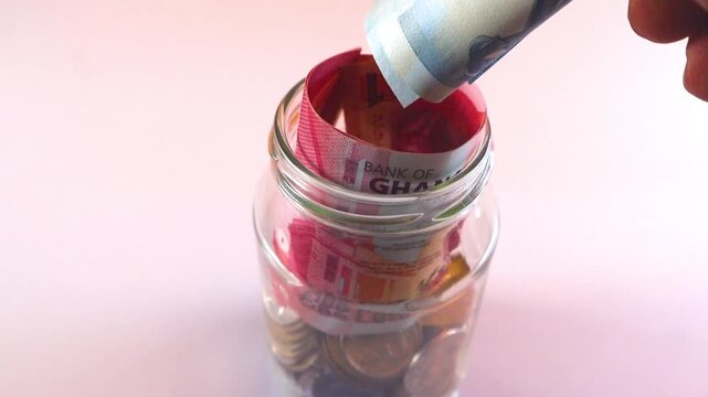 A hand places rolled Ghanaian Cedi notes (GH₵1 and GH₵5) into a glass jar filled with coins. A clear visual of personal savings, financial planning, and Ghana's national currency.