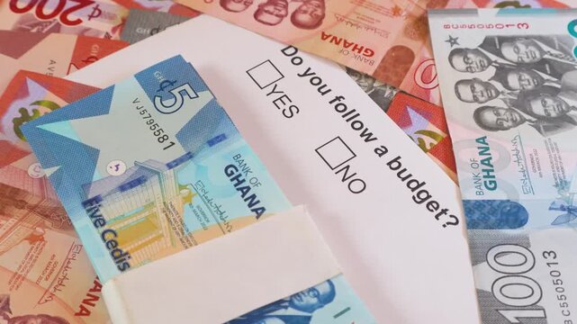 A top-down rotating shot of Ghana Cedi notes surrounding a budget questionnaire. A bundle of 5 Cedi bills sits on the paper, emphasizing themes of personal finance, saving, and monetary accounting.