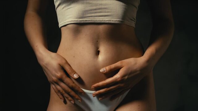 Close-up of a young woman's hands pressing against her bare abdomen and stomach, suggesting period cramps, digestion issues, or body positivity in a dark cinematic lighting setup.