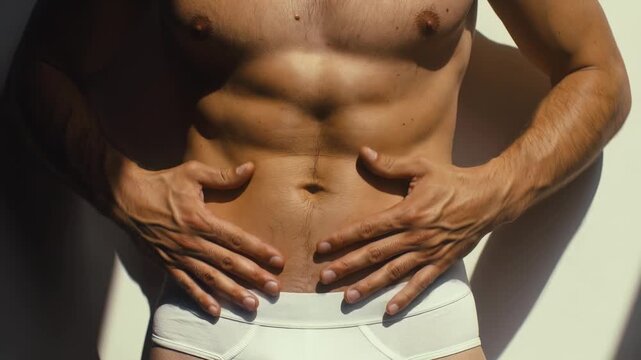 Close-up of a muscular man's fit abdomen as he gently touches his stomach, wearing white underwear under bright sunlight with dramatic shadows, showcasing physical fitness.