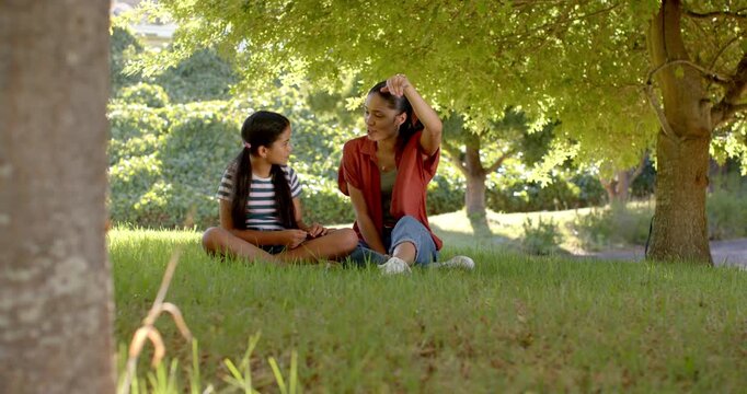 Smiling mother starting conversation guiding daughter in striped top using tablet under park tree