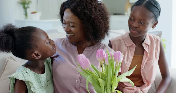 Holding bouquet, mother wearing mauve blouse hugging daughters on sofa at home, sharing pink tulips