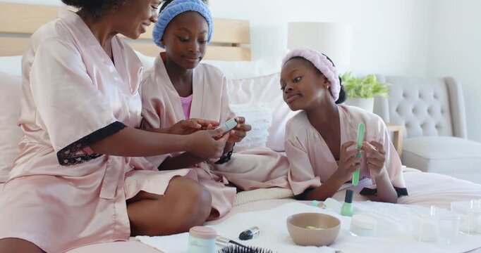 Showing woman and two girls in pink robes passing bottle on bed, pampering with nail file