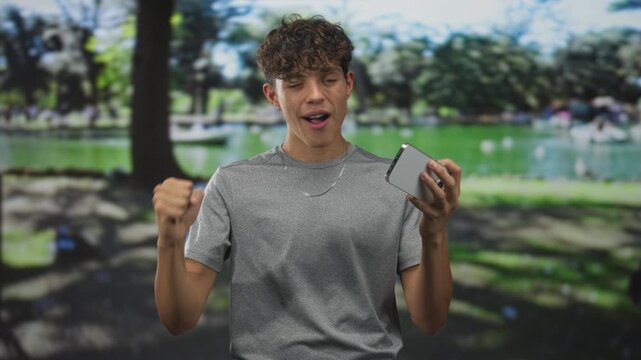 Teenage boy holding smartphone and pumping fist in forest near a pond, smiling broadly at the screen while wearing a gray tee and chain necklace; excitement triumph.