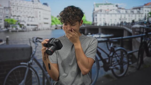 Young man holding dslr camera, hand to mouth gesture on a street beside canal and parked bicycles; surprise curiosity discovery.