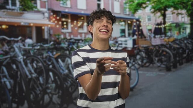 Teen man pointing fingers and smiling, hands visible, in front of parked bicycles on a street; playful joy.