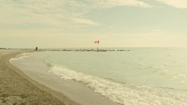 Dynamic waves break on Kew Beach, Toronto as a person stands with a dog on the shoreline. A Canadian flag blows in the wind on a bright March afternoon.