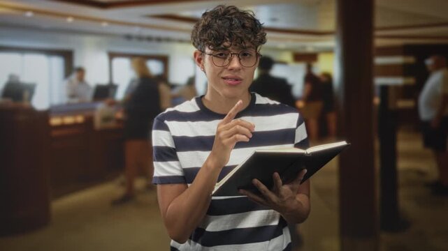 Teenage boy holding book, finger to lips for silence inside building reception area wearing glasses and striped shirt; concentration curiosity.
