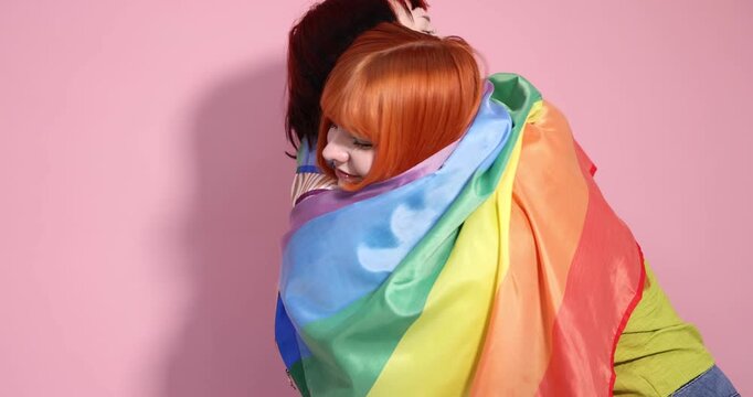LGBT concept. Lovely lesbian couple with rainbow flag hugging each other on pink background. Camera moving forward