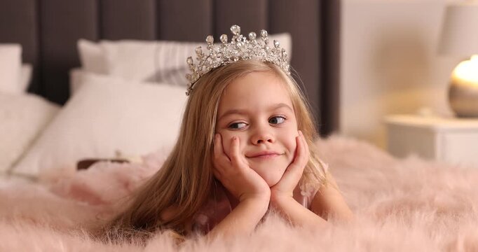 Beautiful little girl with crown dreaming about becoming queen on bed at home