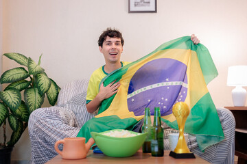 Torcedor brasileiro assistindo a um campeonato internacional de futebol pela TV em casa. Vestindo a camisa da seleção brasileira, ele torce com empolgação no sofá enquanto segura a bandeira do Brasil. © Marcio