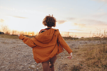 Naklejka premium A woman wearing an orange jacket walks on a rocky path during sunset. The woman enjoys nature and freedom in the open landscape with a clear sky and warm light.