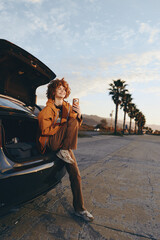 Naklejka premium Young woman with curly hair sitting in car trunk smiling, holding smartphone, wearing casual clothes, outdoor lifestyle, palm trees in background, warm sunlight, relaxed mood.