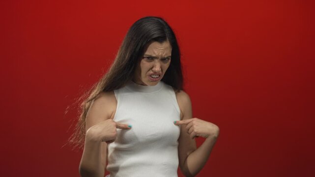 Young hispanic woman in white sleeveless top points both index fingers to chest in studio with red backdrop, furrowed brow and open mouth; skepticism.