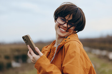 Smiling woman lifestyle wearing glasses and orange jacket holds smartphone outdoors in natural...