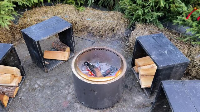 Cozy outdoor winter campfire scene featuring a sleek stainless steel smokeless fire pit surrounded by rustic black wooden crates filled with split logs and golden hay bales under evergreen trees
