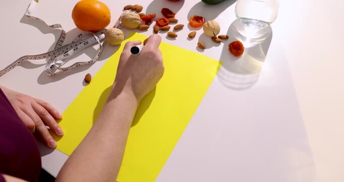 Woman writing diet plan on yellow notepad with healthy foods. Hand planning nutrition menu surrounded by orange nuts and vegetables. Weight control and balanced eating concept. video footage