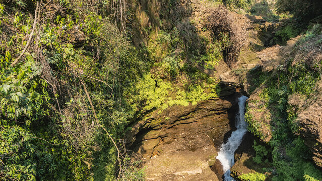 A picturesque waterfall. A stream of white water flows through the gorge between the rocks. Green vegetation on steep slopes. Sunny day. Nepal. Pokhara. Davis Falls.  Paatal Ko Chango. 
