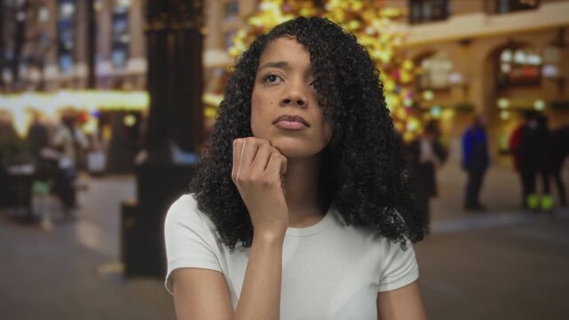 African american woman touches chin on city street beside illuminated christmas tree lights; reflection.