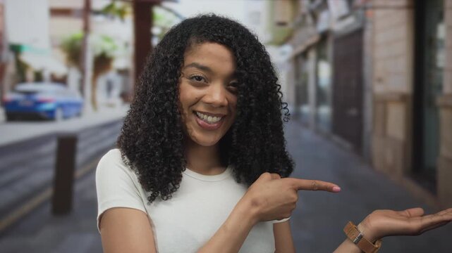 Woman giving thumbs up on a city street amid blurred storefronts and parked car; approval happiness.