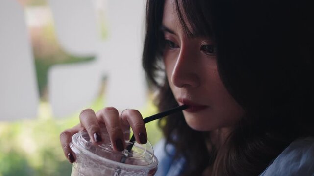 close-up Young Asian woman drinking cold drink through straw relaxing near window quietly