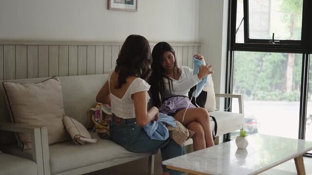 Two young Asian girls sitting on a sofa in a large living room share and try on clothes they have just bought