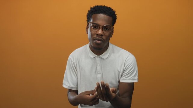 Young african american man rubbing his fingers in money gesture in orange studio setting for financial plea; desire wealth.