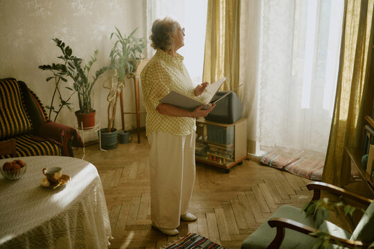 Senior Caucasian woman standing in living room holding open photo album and looking toward window, sunlight streaming through sheer curtains, indoor plants and vintage decor visible in background
