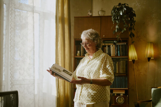 Senior Caucasian woman standing in living room holding open photo album, bookshelf and houseplant in background, natural light streaming through window illuminating scene