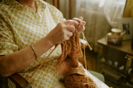 Senior Caucasian woman sitting indoors knitting with yarn and needles, hands holding partially completed project, wearing patterned shirt, natural light illuminating workspace