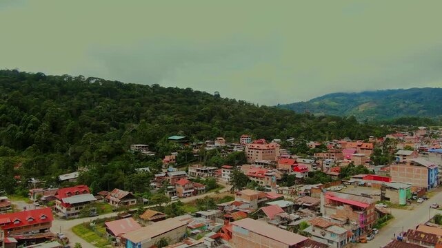 Drone orbits the city of Villa Rica, Peru, nestled within the high jungle of Chanchamayo, showing dense green mountain vegetation under a cloudy sky.