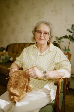 Portrait of senior Caucasian woman sitting in armchair knitting with brown yarn, wearing eyeglasses and earrings, looking directly at camera with calm facial expression