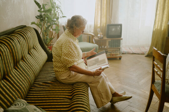 Senior Caucasian woman sitting on sofa holding photo album, looking at old photographs in vintage living room with wooden floor, television and houseplants visible in background