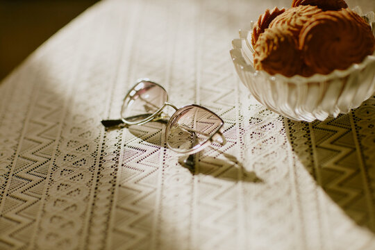 Eyeglasses resting on lace tablecloth near bowl filled with round cookies, sunlight casting shadows across surface, highlighting everyday objects in domestic setting