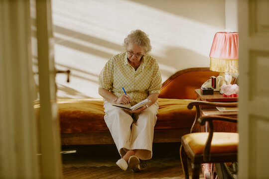 Senior Caucasian woman sitting on sofa writing in notebook with pen, wearing eyeglasses, concentrating on journaling or making notes, surrounded by home decor, natural light streaming in