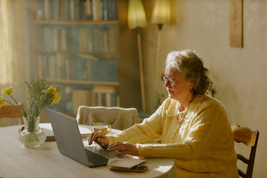 Senior Caucasian woman sitting at table using laptop, typing on keyboard with focused expression, bookshelf and flowers in background, eyeglasses resting on face, engaging with technology