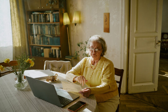 Senior Caucasian woman sitting at table using laptop while holding document, wearing eyeglasses, surrounded by papers and cup, working or studying in home environment