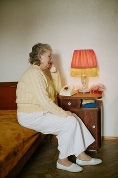 Senior Caucasian woman sitting on bed talking on vintage rotary phone, wearing glasses and earrings, lamp and small objects on wooden nightstand, looking focused during conversation