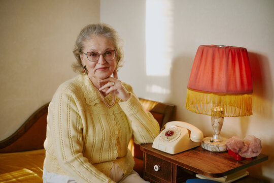 Portrait of senior Caucasian woman sitting beside vintage rotary phone on wooden nightstand, wearing eyeglasses and jewelry, resting hand on chin while looking at camera