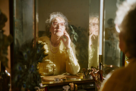 Senior Caucasian woman sitting at vanity table touching face and looking at reflection in mirror, wearing eyeglasses and jewelry, surrounded by personal grooming items and soft natural light