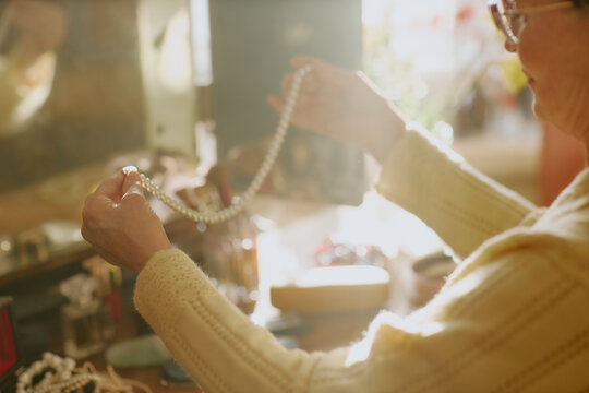 Senior Caucasian woman holding pearl necklace with both hands, examining jewelry at dressing table, sunlight illuminating hands and accessories, only upper body and hands visible