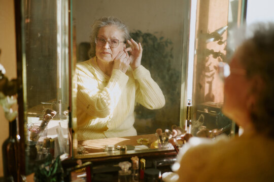 Senior Caucasian woman adjusting earring while standing in front of mirror, wearing glasses, looking at own reflection, hands raised near ear, surrounded by personal care items