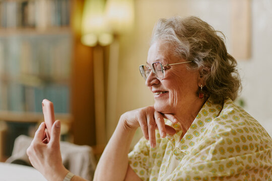 Senior Caucasian woman smiling while holding smartphone, sitting indoors and looking at device screen, wearing eyeglasses and patterned shirt, appearing engaged in video call