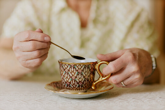 Senior Caucasian woman holding ornate teacup with one hand while stirring beverage with small spoon using other hand, sitting at table, only hands and part of torso visible